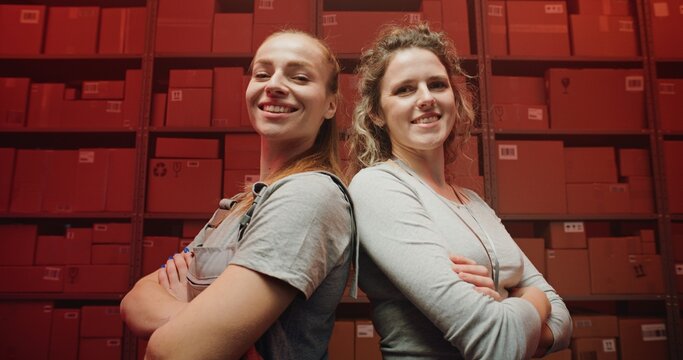 Two Female Warehouse Workers Standing with Crossing Arms, Smiling, Looking at Camera. Postal Delivery Service, Distribution Center with Shelves full of Cardboard Boxes. Red Neon Lighting. Dolly Shot.
