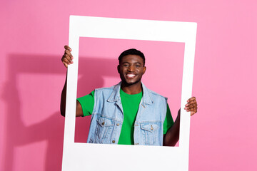 Smiling young man in casual fashion against a pink background holds a large white frame, showcasing...