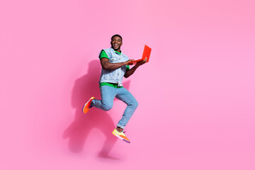 Energetic young man with black hair joyfully jumps while holding a laptop against a pink backdrop...