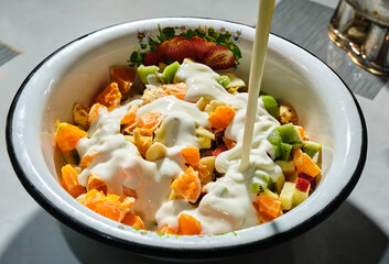 Delicious fruit salad with pieces of kiwi, orange, and apple, drizzled with yogurt, in a white bowl with a floral pattern.
