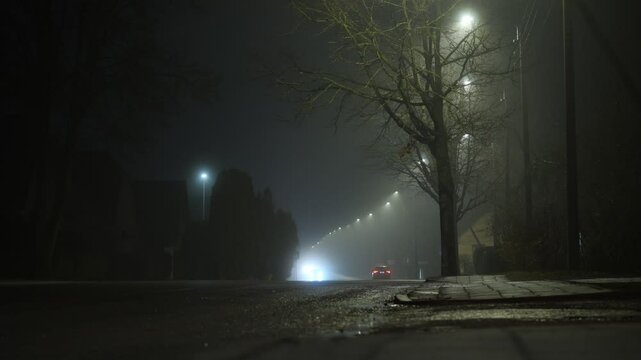 Small, misty road at night with headlights cutting through the fog.