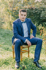 Obraz premium A man in a suit and bow tie sits on a wooden chair in a grassy field. The scene is serene and peaceful, with the man looking off into the distance. The combination of the man's formal attire