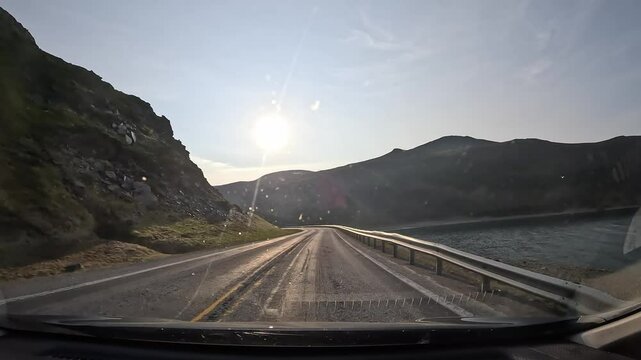 Driving through tundra on the E69 road in northern Norway. Rocky hills with grass surround the route. A sea bay appears on the right. The sun shines ahead as the car enters the Nordkapp tunnel.