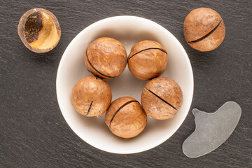 Macadamia nuts in shell with ceramic saucer on slate stone, top view.