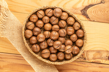 Macadamia nuts in shell with straw plate on wooden table, top view.