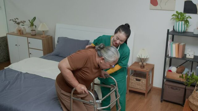 High angle shot of elderly woman with walker getting up from bed with assistance of Asian female caregiver visiting patient at home