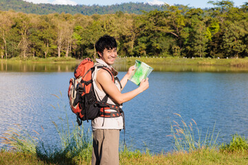 An Asian young man traveler with an old paper map smiling and eyes contact look at camera beside a lake during sunny day
