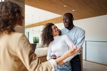 Female real estate professional in business attire consults with diverse couple using tablet device during property viewing meeting in modern office.