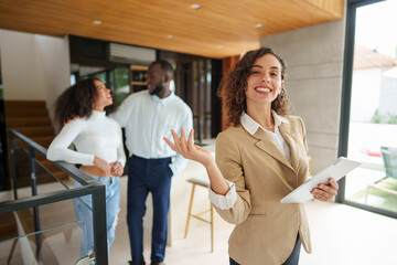 Female real estate professional in beige suit presents property details using tablet while diverse couple explores modern luxury home interior.