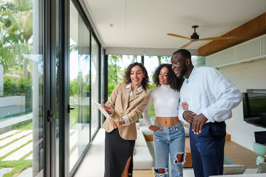 Professional female real estate agent in blazer presents modern glass-walled property to diverse couple during daytime viewing tour of contemporary residential space. - Powered by Adobe
