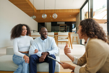 Professional real estate agent in business attire conducts consultation with diverse couple in modern living room setting using digital tools for property discussion.