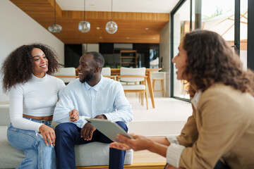 Female real estate professional in business attire reviews digital tablet documentation with diverse couple during property consultation in contemporary indoor setting.