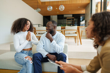 Female real estate professional in business attire consults with diverse couple using digital tablet during property discussion in modern living space.