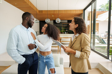Female real estate professional in tan blazer discusses property details with diverse couple in...