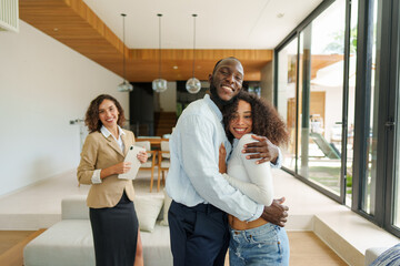 Young diverse couple shares celebratory moment with female real estate agent in modern residential property interior wearing business attire during successful home purchase completion.