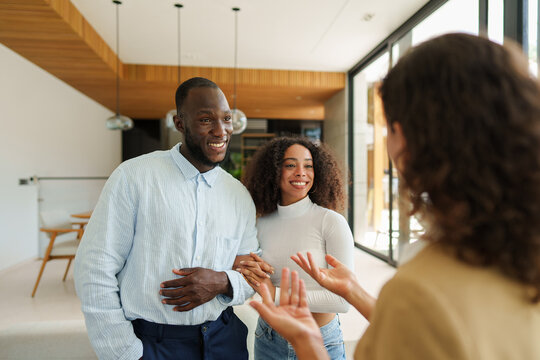 Diverse couple tours modern luxury home with real estate agent, sharing excited conversation during property viewing in bright contemporary interior setting.