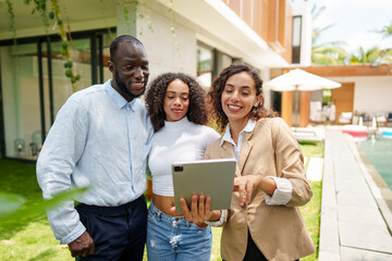 Professional real estate agent in business attire presents property details using tablet to diverse clients during outdoor residential property consultation.