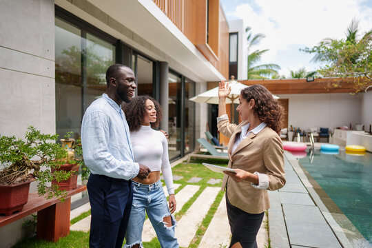 Female real estate agent in business attire presents modern luxury property with pool to diverse couple during outdoor property tour consultation. - Powered by Adobe