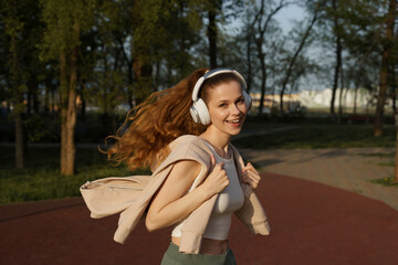 a young beautiful woman in sports clothes is running in park, a photo in motion, woman is smiling with wireless headphones, curly hair in ponytail