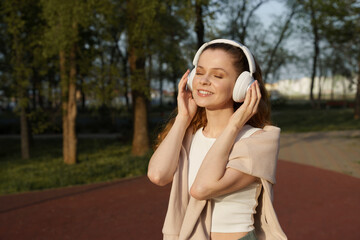 A young woman with headphones listening to music in a park, wearing a white top, with trees in the background. Concept of relaxation and outdoor leisure.