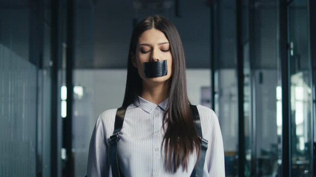 Young professional woman standing in an office corridor with her mouth taped shut by a man's hands, symbolizing censorship or workplace inequality. Concept of oppression and silencing