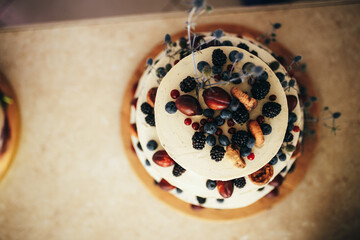Tiered rustic cake topped with fresh blackberries, blueberries, red currants, chestnuts, and nuts, accented with natural flowers on a wooden base. Wedding Cake with Fresh Berries.