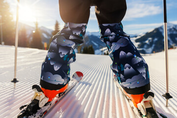 Scenic back POV view kid beginner skier close-up legs downhill start line rows freshly prepared groomed ski slope piste bright warm morning sunrise sky background.  mountain landscape winter resort