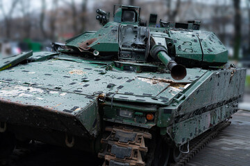 Military tank parked on city street surrounded by trees and urban buildings during overcast weather
