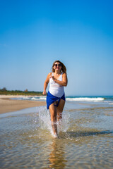 Fototapeta premium Beautiful middle-aged woman running on sandy beach in summertime. Physical activity on beach. Front view