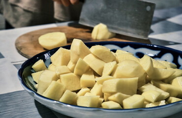 A close-up of the process of cutting potatoes on a wooden board. In the center of the frame, there's a knife and a potato, with a bowl of sliced potatoes in the foreground.