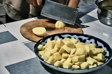 A close-up of the process of cutting potatoes on a wooden board. In the center of the frame, there's a knife and a potato, with a bowl of sliced potatoes in the foreground.