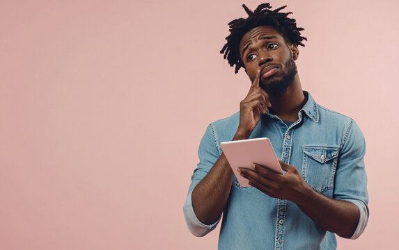 Confident man in denim shirt holding a tablet, standing with a thoughtful expression, against a soft pink background, reflecting a moment of contemplation.

