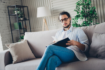 Obraz premium Handsome young man relaxing at home on a sofa while reading a book in a comfortable and well-lit interior