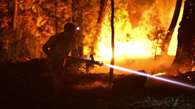 Nighttime forest fire engulfs mountain slopes with heavy smoke. Firefighters with hoses and axes battle large flames, capturing a dramatic view of this natural disaster in tropical forests.