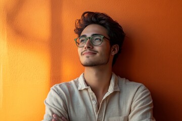 A young Latino man with glasses stands against a vibrant orange wall, looking thoughtfully upwards with a slight smile