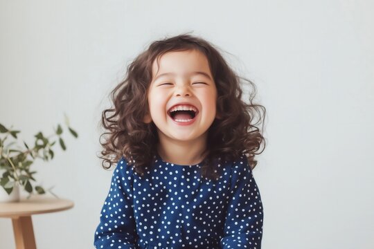 six year old girl with medium brown skin and curly pigtails, wearing a blue polka-dot dress, sitting on a stool and laughing, set against a pastel pink background.