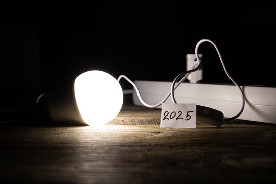 An energy saving light bulb is on a wooden floor near a socket and a card with the number 2025 - Powered by Adobe