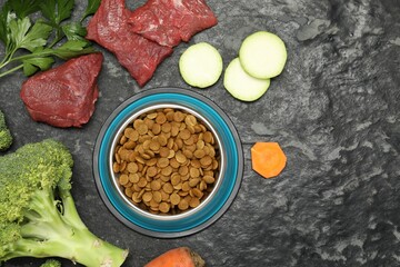Dry pet food in feeding bowl and natural products on dark textured table, flat lay. Space for text