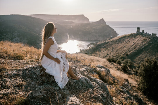A woman in a white dress sits on a rock overlooking a body of water. The scene is serene and peaceful, with the woman enjoying the view and the calmness of the surroundings. - Powered by Adobe