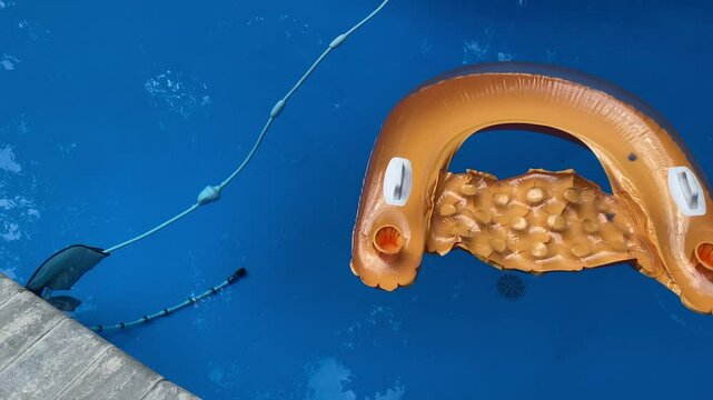 A video of a poolside view showing two gold inflatable pool chair floats with cup holders drifting in the water as an electric 360-pressure side pool cleaner glides by beneath them.