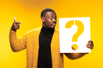 Thoughtful man holding a question mark sign on a yellow background, wearing a cozy brown sweater,...