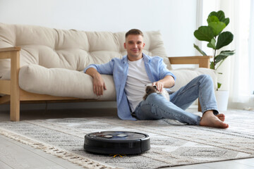 Young man with cute cat at home, focus on robotic vacuum cleaner