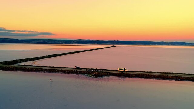 An aerial view of a long road crossing a vast salt lake at sunset, with vibrant orange and pink hues reflecting on the calm water, creating a serene and surreal atmosphere.