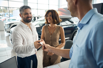 Fototapeta premium Happy man with woman putting car key from salesman at the dealership showroom