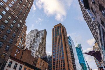 Looking up at New York City skyscrapers with diverse architectural styles against blue sky and clouds.