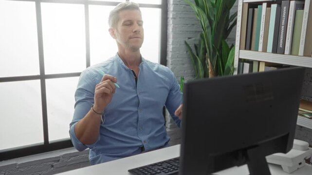 Young man sings using pen as a microphone in a modern office setting with bookshelves and computer nearby, expressing joy and creativity during work.