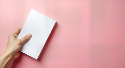 Female hand holds blank white book cover against pastel pink background. Design mockup perfect for book covers paper products. Image offers plenty of space for custom design. Subtle aesthetic creates