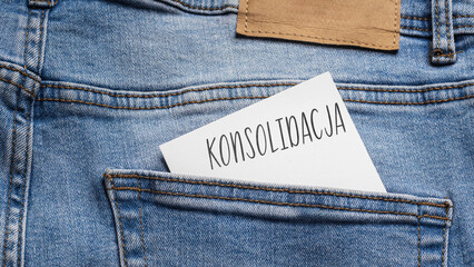 White card with a handwritten inscription "Konsolidacja", inserted into the pocket of blue pants jeasnow (selective focus), translation: Consolidation