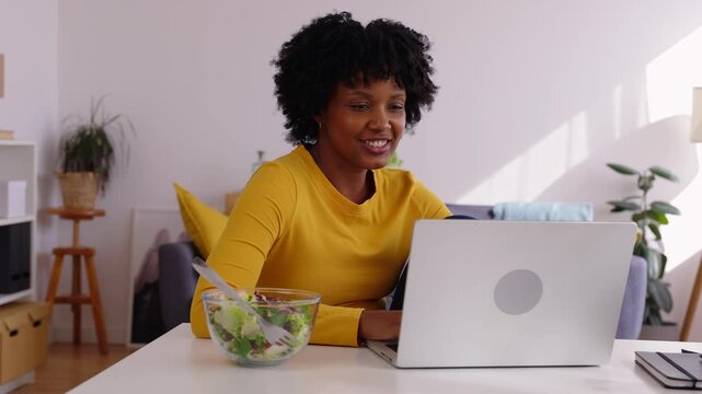 Happy young woman working on laptop computer from home and eating a healthy salad during her lunch break. Technology and healthy lifestyle concept.
