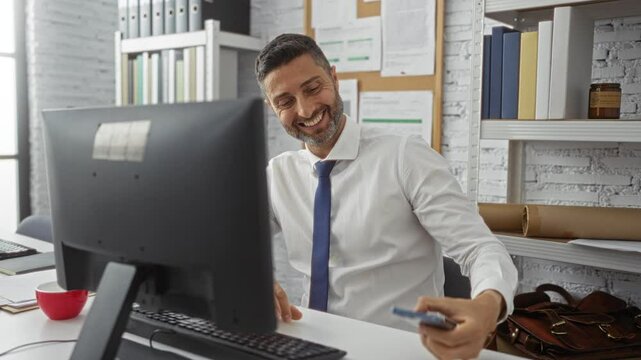 Smiling man in office making video call on smartphone while sitting at desk with computer during workday ambiance.